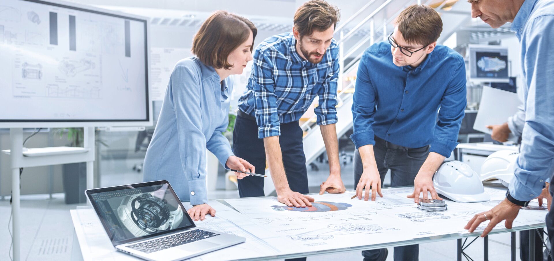 © AdobeStock_292341232_Gorodenkoff (In the Industrial Engineering Facility: Diverse Group of Engineers and Technicians on a Meeting Gather Around Table with Engine Design Technical Drafts, Have Discussion, Analyse Technology)